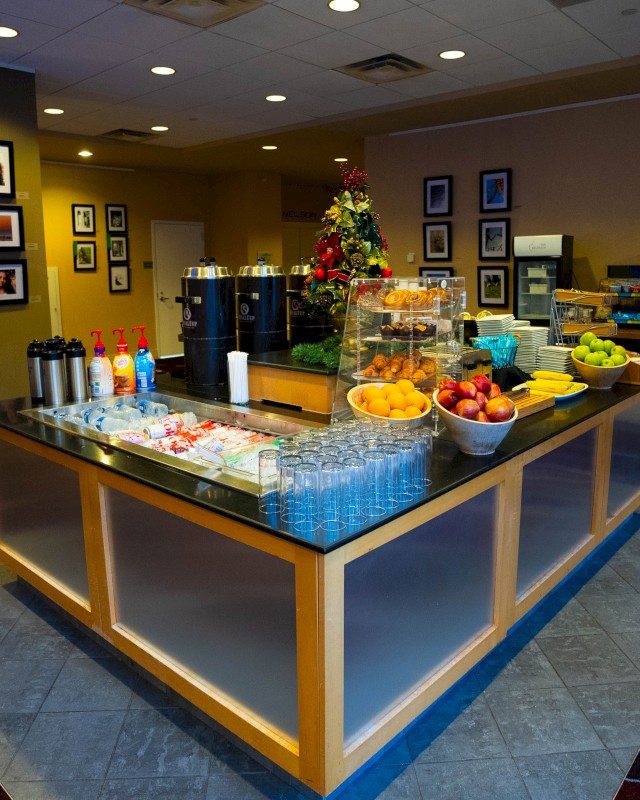 A hotel breakfast buffet features fruits, beverages, pastries, and condiments on a counter in a well-lit room with framed pictures.