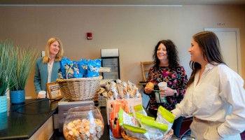 Three people in a cafe, surrounded by snacks and drinks, engaged in conversation while holding food and beverages.