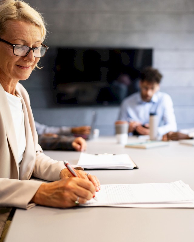 A woman in a blazer writes on a document at a conference table, with three people blurred in the background during a meeting.