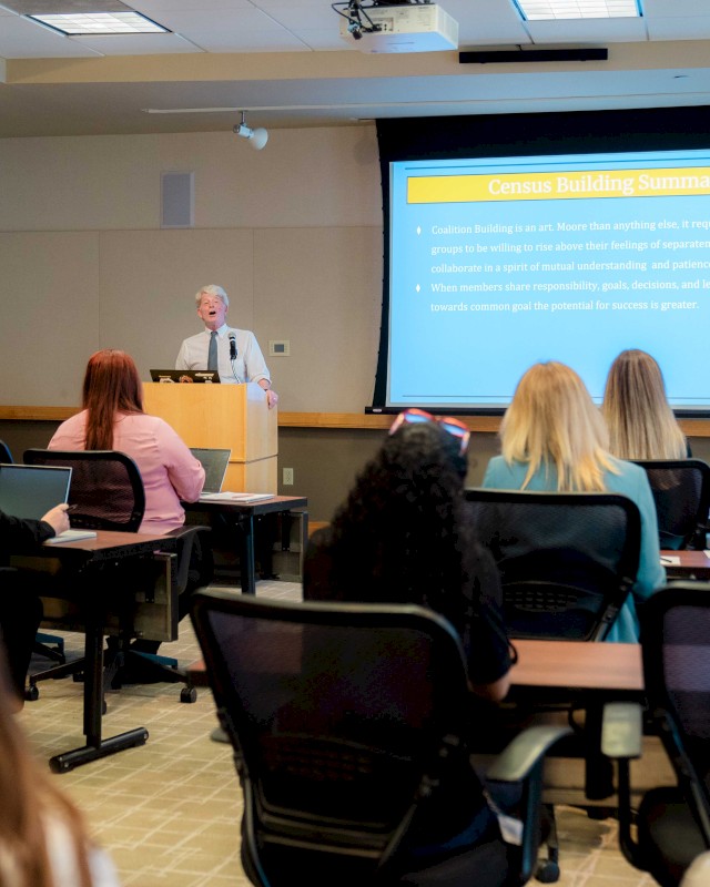A group of people in a classroom setting watching a presentation titled 
