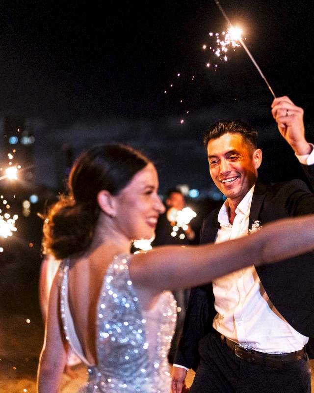 A couple is joyfully celebrating with sparklers at a night event, surrounded by smiling guests.