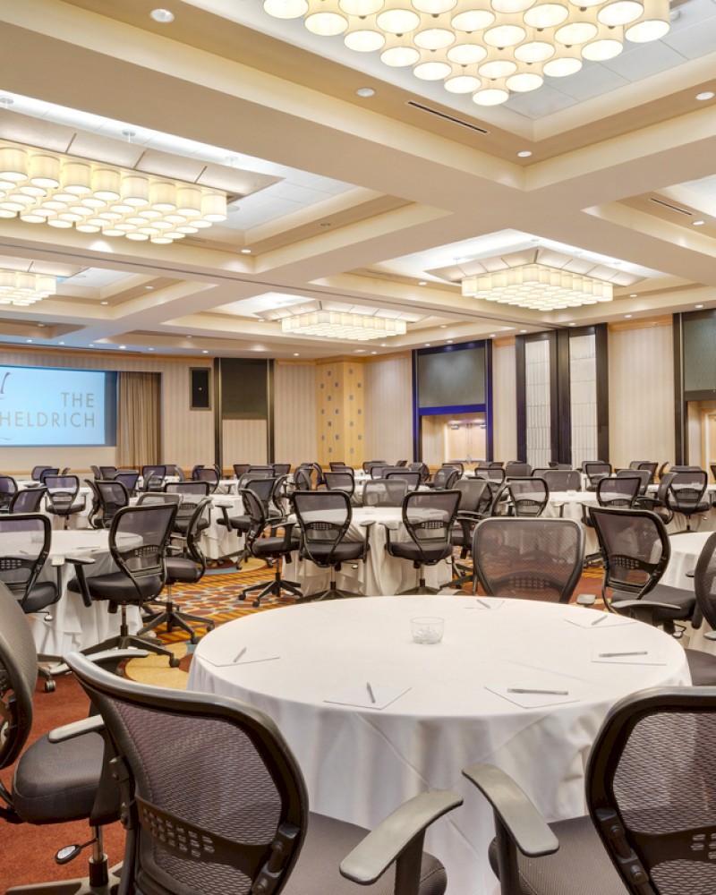 A conference room with round tables, mesh chairs, and a large screen. The ceiling has rectangular light fixtures, and the floor is carpeted.