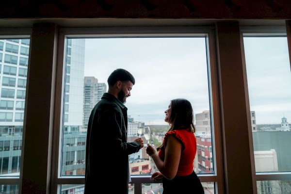 A couple stands by a large window, exchanging smiles, with a cityscape view in the background.