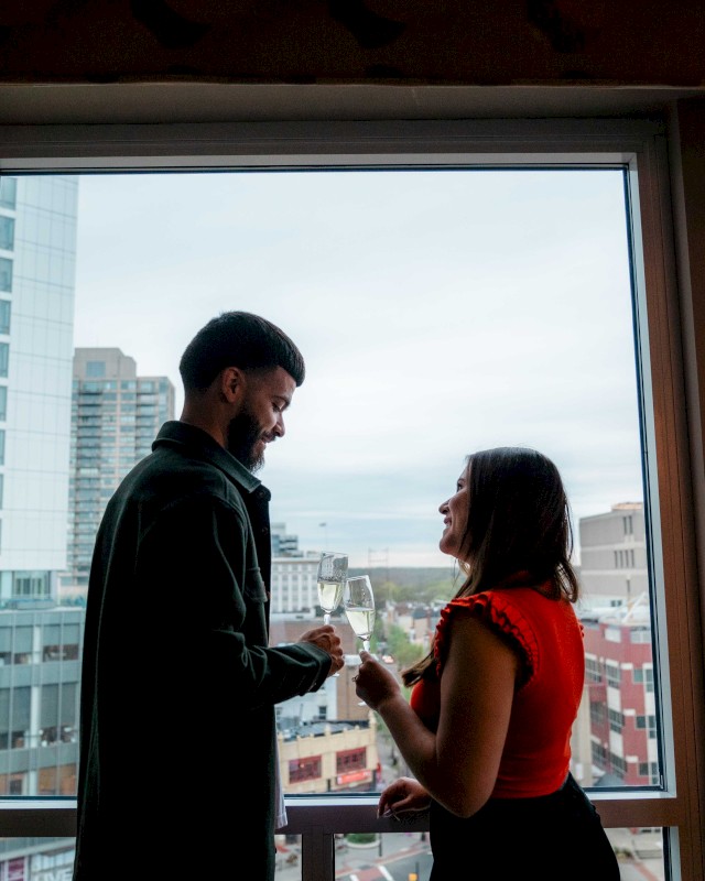 A couple stands by a large window, exchanging smiles, with a cityscape view in the background.