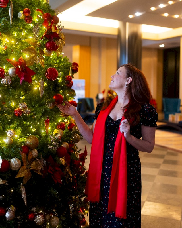 A person with a red scarf admires a decorated Christmas tree with red and gold ornaments in a warmly lit indoor setting.