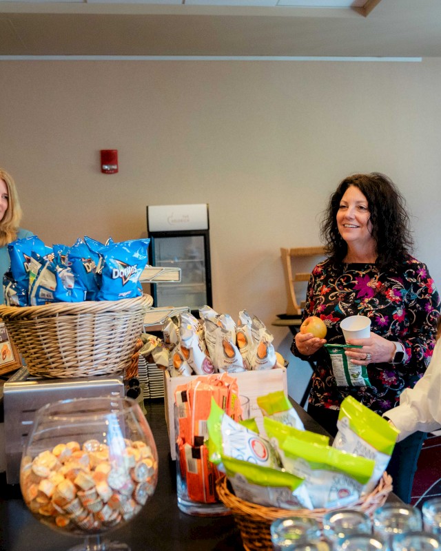 Three women are chatting at a snack counter with bags of chips, baskets, and a jar of small treats on display.