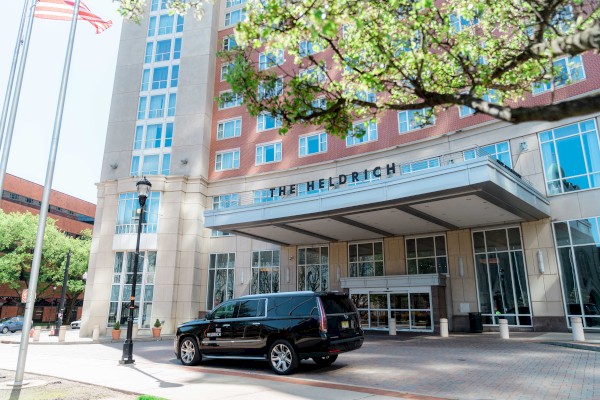 The image shows the entrance of The Heldrich hotel, featuring a large building facade, an SUV parked in front, and nearby trees and flagpoles.