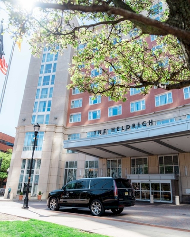 A black SUV is parked in front of a modern hotel entrance, with a tree in bloom and a sunlit building labeled “THE HELDRICH.”