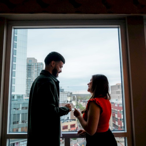 A couple stands by a large window in a high-rise, sharing a moment and possibly exchanging rings, with a cityscape view behind them.