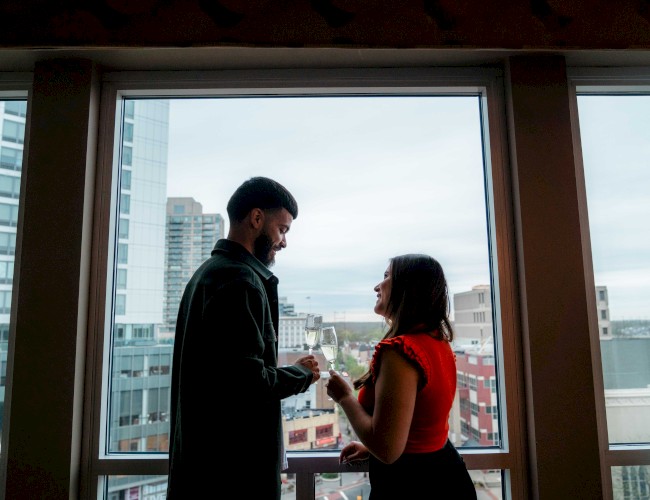 A couple stands by a large window in a high-rise, sharing a moment and possibly exchanging rings, with a cityscape view behind them.