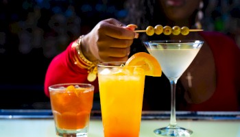 A person decorates a martini with olives on a stick, alongside two other cocktails garnished with orange slices.
