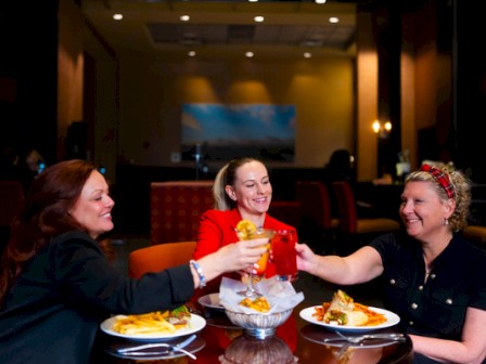 Three friends toast at a restaurant table, smiling with drinks and plates of food in front of them, sharing a cheerful moment together.