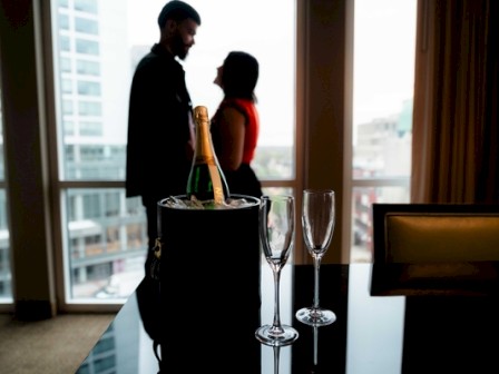 Two people stand close and face each other in a dimly lit room, with a champagne bottle, two fluted glasses, and a bucket on a table in the foreground, suggesting a romantic moment.