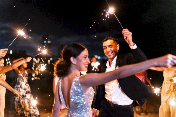 A couple dances at a festive night party with sparklers and other guests in elegant attire around them, celebration vibes all around.