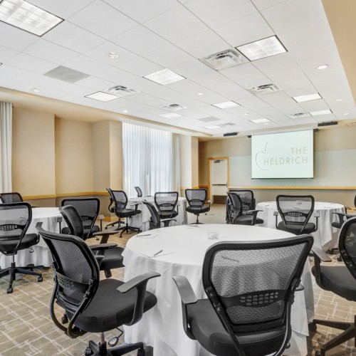 The image shows a conference room with round tables, black mesh chairs, and a presentation screen on the wall.
