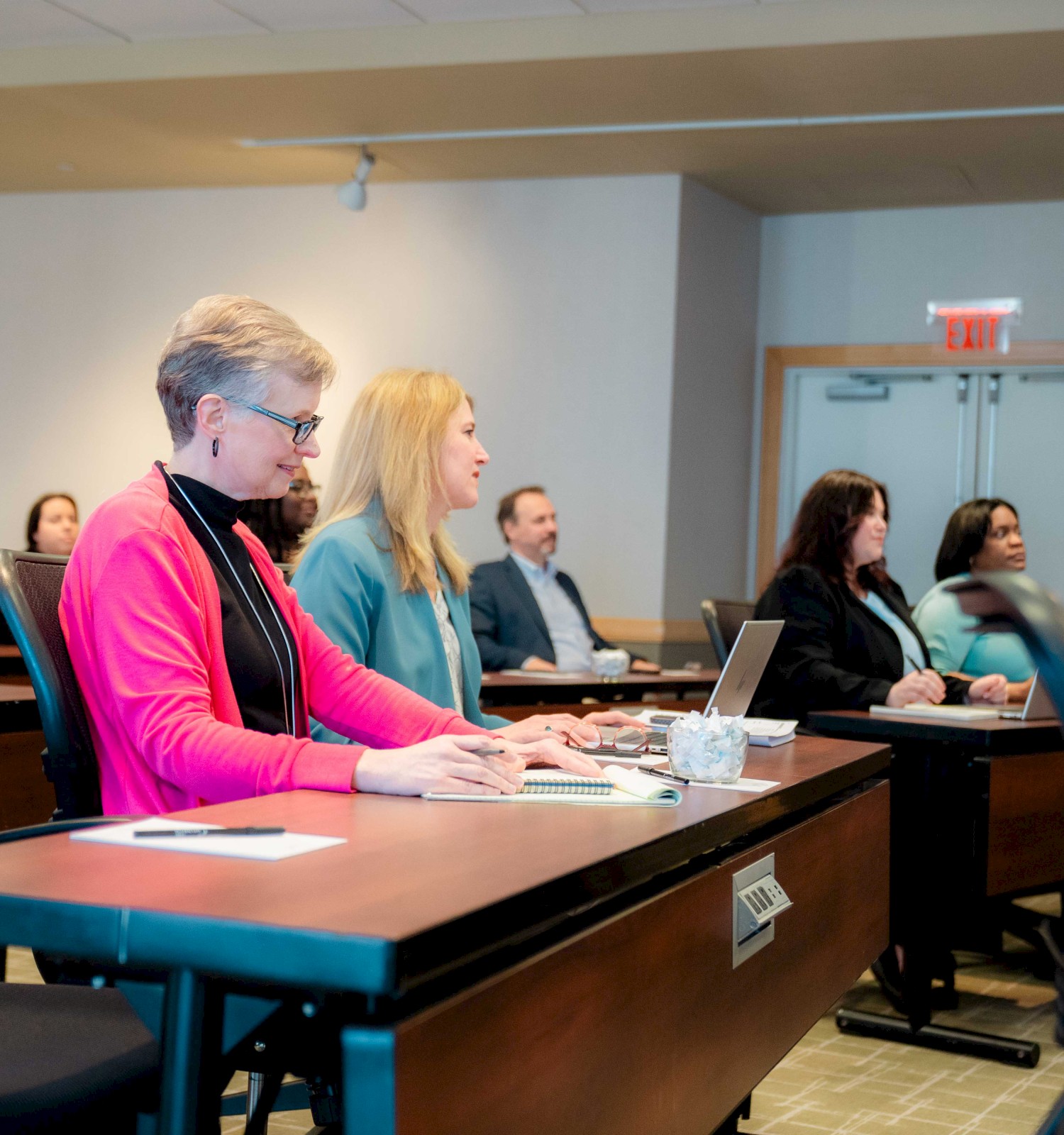 People sit in a classroom or meeting room, listening at desks with laptops and papers, as a presenter or instructor leads the session.