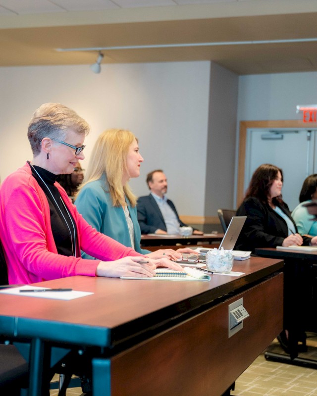 A classroom or meeting room with adults seated at desks, listening attentively; a woman in a pink jacket sits near the foreground, papers and a water cup on the desk.