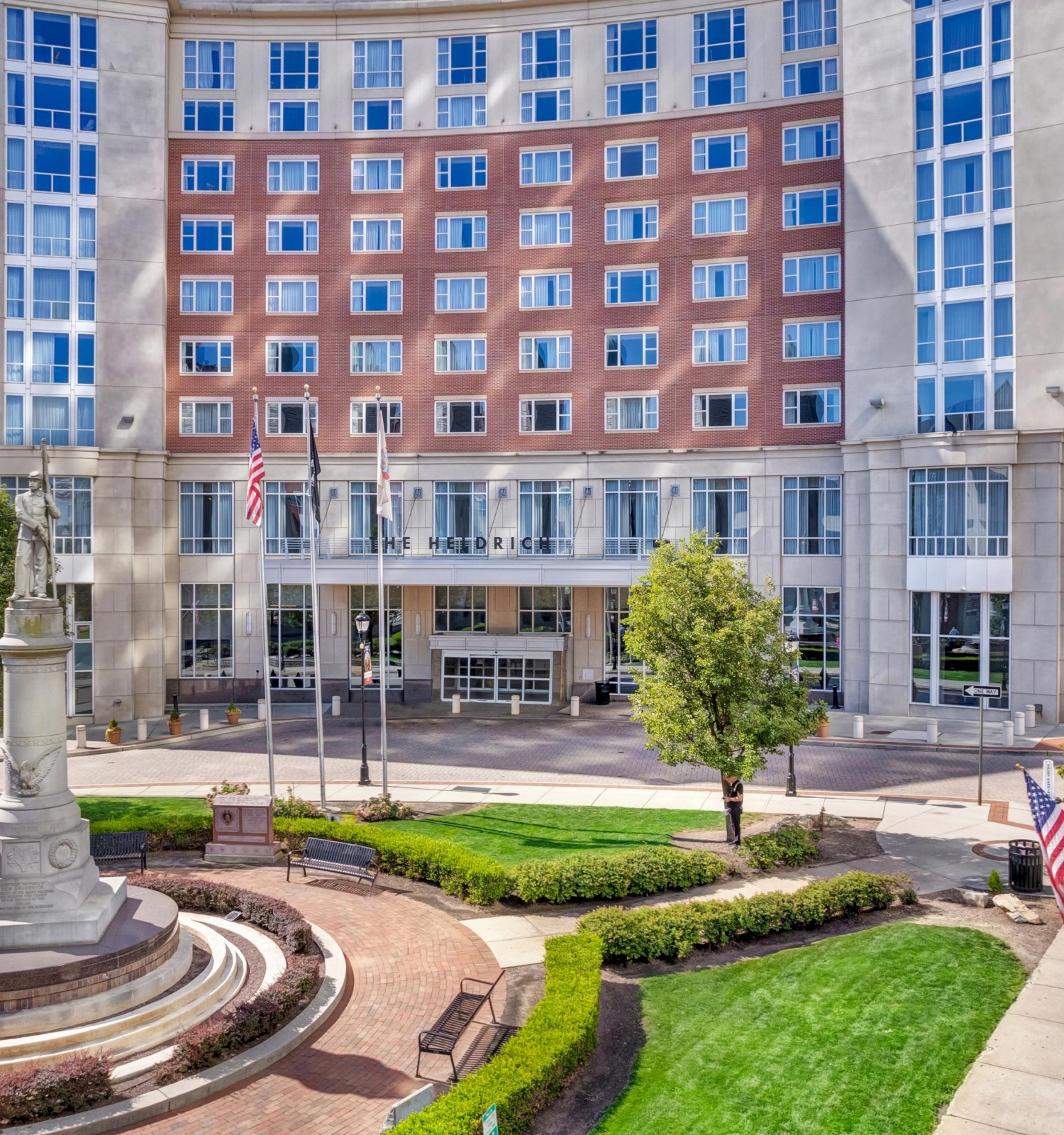 A large, multi-story brick and glass building with a circular plaza, green lawns, flags, and a statue in front, bathed in daytime light, welcoming and civic.