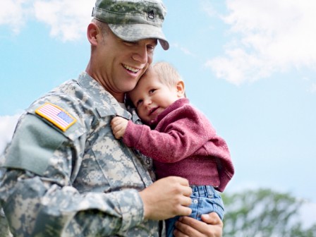 A joyful soldier in uniform holds a smiling child close, a tender moment of care between parent and little one, outdoors on a sunny day.