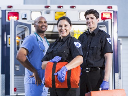 Three healthcare professionals stand smiling in front of an ambulance; a nurse/paramedic holds an orange bag, flanked by two officers.