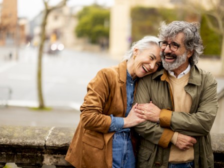 Two smiling older adults lean on a railing, hugging happily on a city street, sharing warmth and companionship as they enjoy a sunny day.