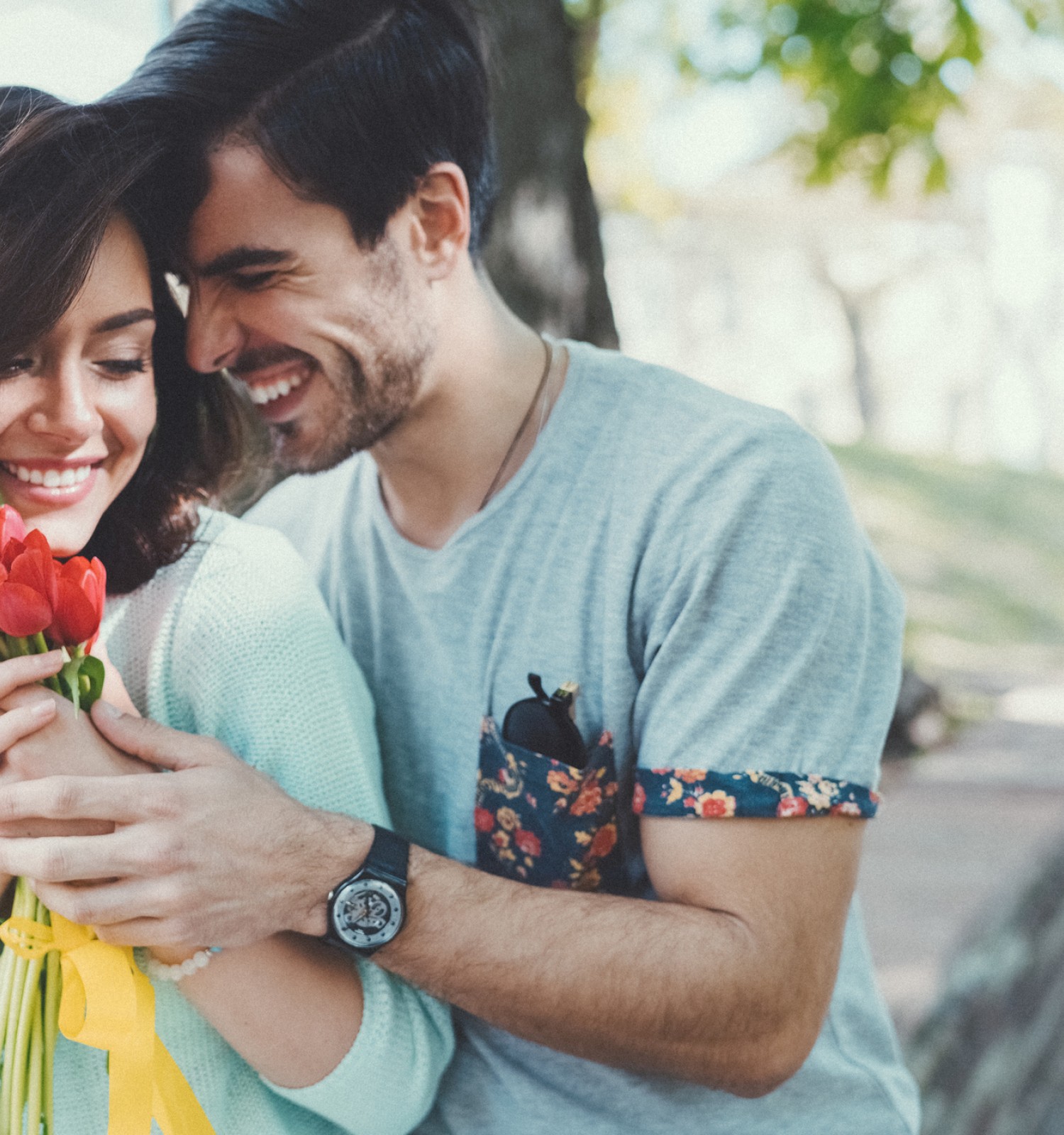 A couple smiles while a man hand-holds a bouquet of red flowers for his partner in a sunny park.
