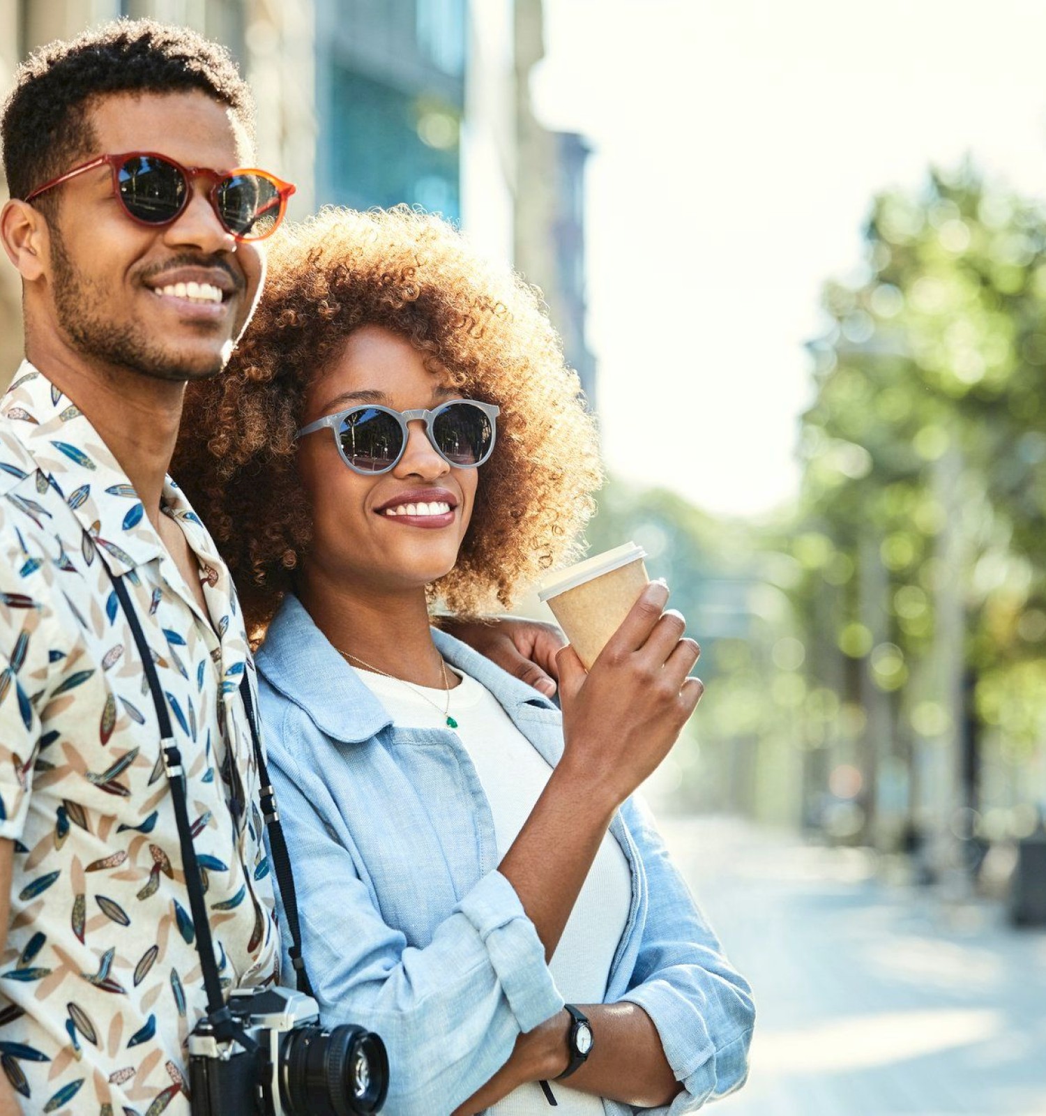 Two stylish friends stand on a sunny street, smiling at the camera, one holding a coffee cup and wearing sunglasses.