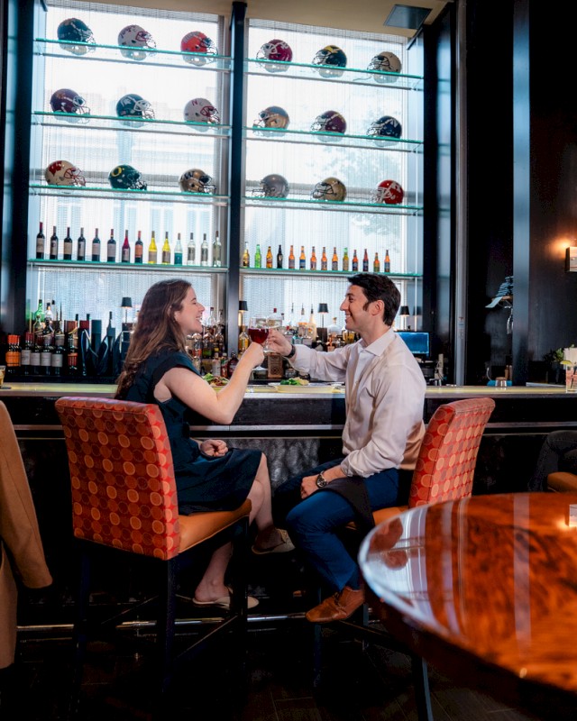 A couple toasting with drinks at a bar, with sports helmets and bottles displayed on shelves behind them.