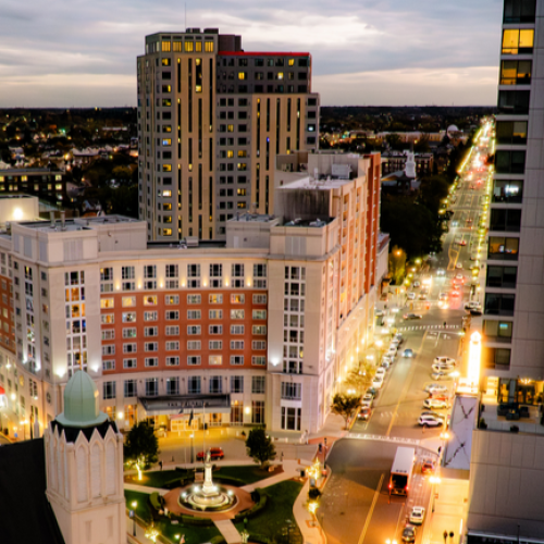 A cityscape at dusk with tall buildings, a lit street, and a roundabout featuring a statue fountain, seen from a high vantage point.