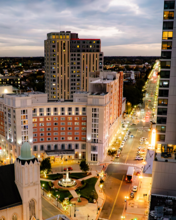 A cityscape at dusk with tall buildings, a lit street, and a roundabout featuring a statue fountain, seen from a high vantage point.