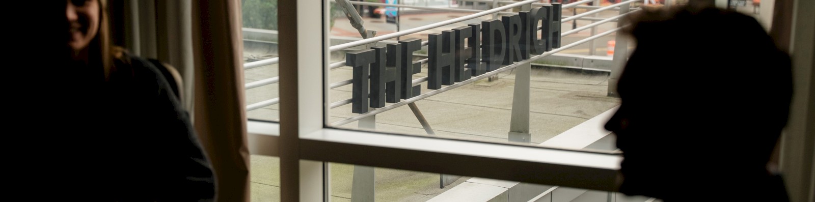 Two people sit by a window in a dim room, shadowed faces, a sign reading 