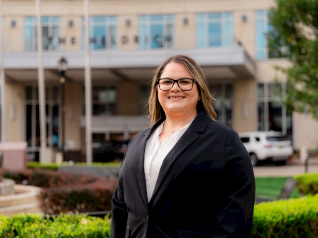 A professional woman in a black blazer stands smiling outside a modern building, with greenery and a driveway in the background.