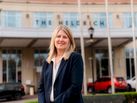 A confident woman stands outside a modern building labeled “THE HELI(R)ICH,” wearing a navy blazer and white blouse, smiling near cars.