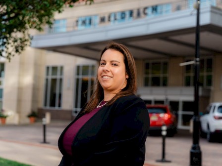 A confident woman in a black blazer and maroon top stands outside a building with a sign and cars in the background.