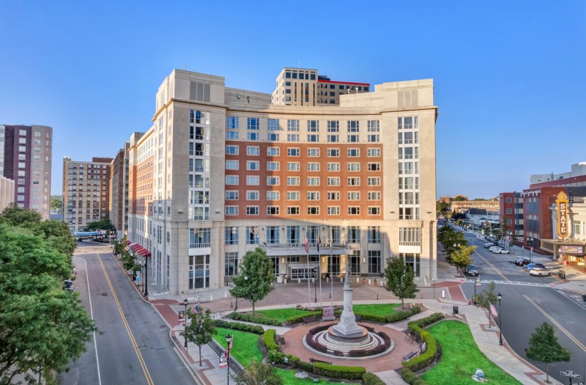 A tall building with a small park and memorial in front, streets, trees, and other buildings on a sunny day in a cityscape.