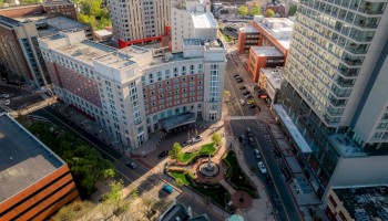 An aerial view of a city block with tall buildings, roads, a small park with a fountain, and various parked cars.