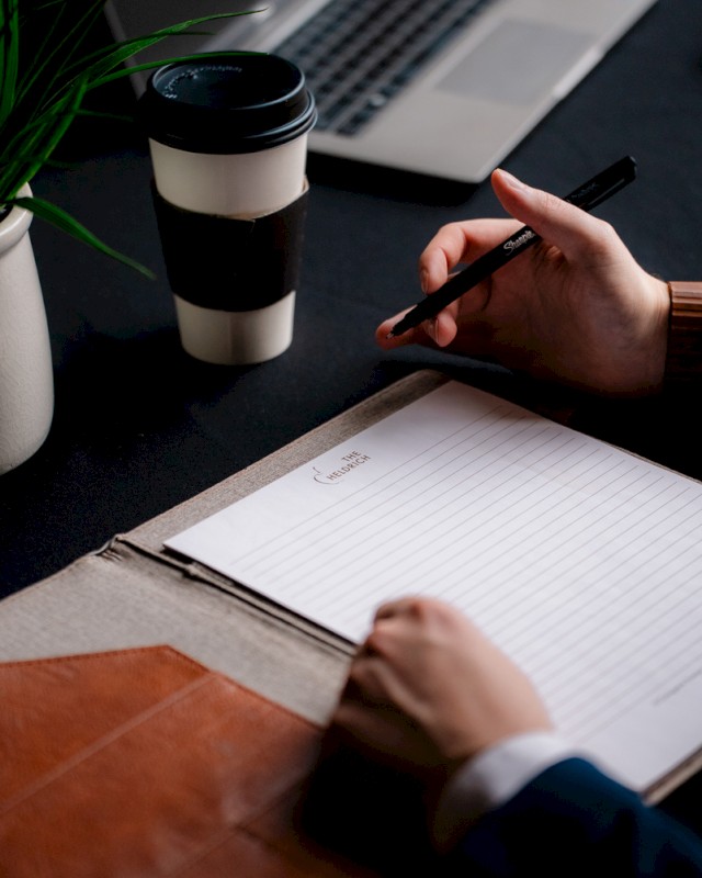 Someone is taking notes in a lined notebook with a laptop and coffee cup nearby, on a dark workspace.