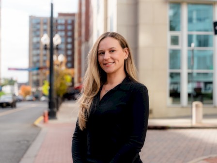 A smiling woman with long blonde hair stands outdoors on a city street with buildings and streetlights in the background.