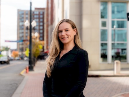 A woman with long blonde hair in a black top smiling outdoors in an urban setting with buildings and street in the background.
