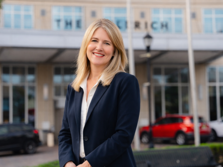 A confident woman in a navy blazer stands smiling outside a building with the word 
