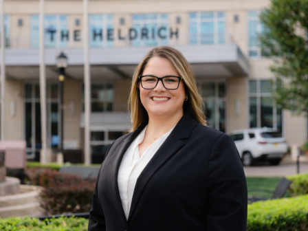 A woman dressed in a black blazer and glasses stands outdoors in front of The Helderich building with greenery and parked cars behind her.