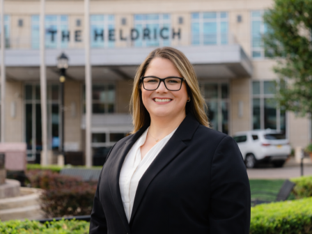 A smiling woman in business attire stands outside The Meldrich building, with trees and parked cars in the background.