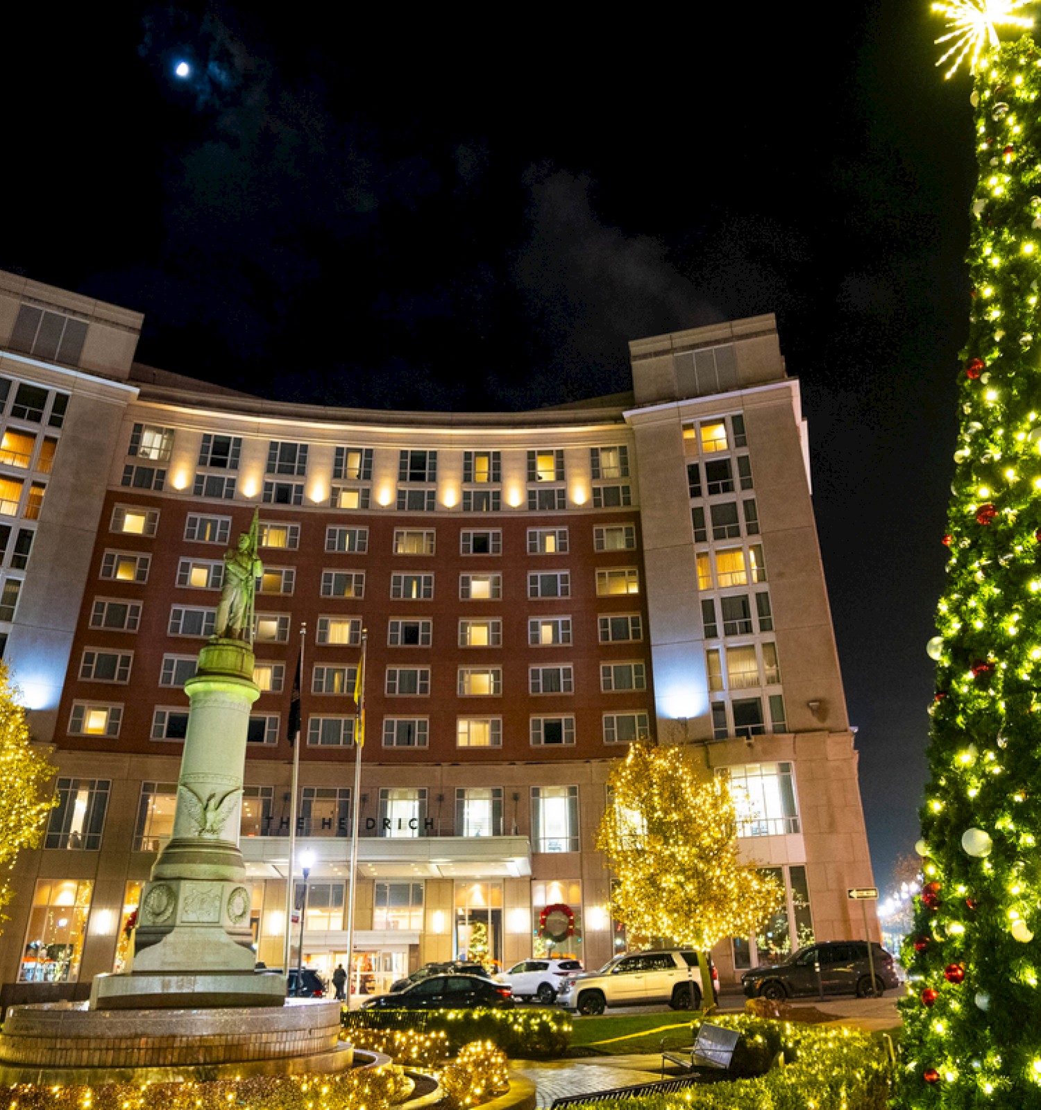 A beautifully decorated Christmas tree and illuminated trees in front of a modern building at night for the holiday season.