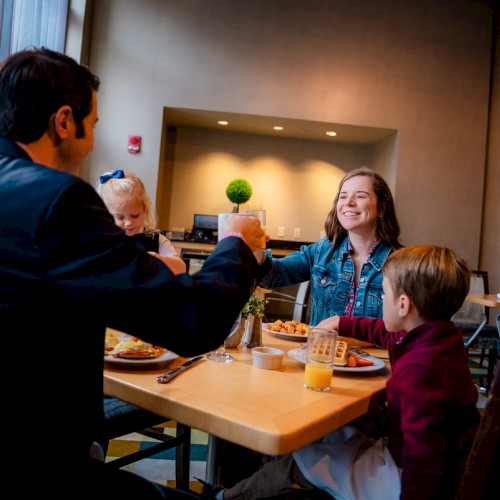 A family enjoys breakfast at a table, sharing a toast with happy smiles in a cozy, well-lit restaurant setting.
