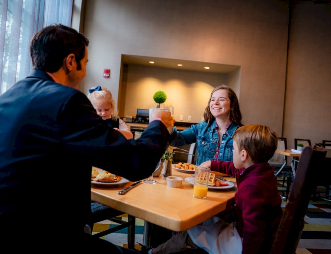 A family enjoys breakfast at a table, sharing a toast with happy smiles in a cozy, well-lit restaurant setting.
