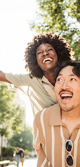 A joyful woman with arms wide open and a man smiling outdoors on a sunny day in a city park or street.