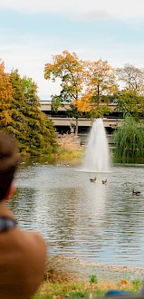 A couple watches ducks on a pond surrounded by autumn trees and a parking garage in the background.