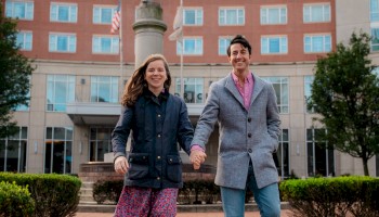 A couple happily walks hand in hand in front of a large hotel or apartment building, holding hands and smiling outdoors.