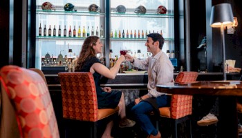 A couple is enjoying drinks at a bar, clinking glasses and smiling in a cozy, modern setting with sports helmets on the shelves.