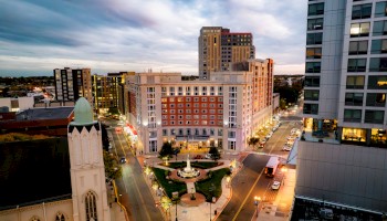 A cityscape with tall buildings, a church, and a park with a fountain at sunset, illuminated by streetlights and city lights.
